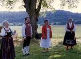 Midsommarfirande vid Långåker Hembygdsgård år 1992. Fyra folkdräktsklädda medlemmar står utomhus. Från vänster: Eivor Bjerrhede (1921 - 2015), sonen Staffan, Gun Flygare och Marianne Jansson.