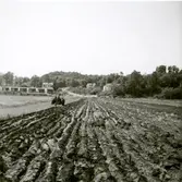 Sven Svensson plöjer med traktor på åkern Kärret, Vommedal 3:2 cirka 1958 - 1959. Till vänster ses Ävavägens radhus. I mitten slingrar sig Bergsjövägen uppför backen.