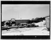 Vägstation B7, Åkersberga. Baksidan av förråds- och verkstadsbyggnaden. Luttunnor upphängda i ställningar. Upplag av bränslefat.