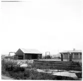 Vägstation D7, Jönåker. Översikt med förrådsbyggnad till vänster och del av garagebyggnaden till höger. Bensinpumpar (drivmedelspumpar) vid lastkaj. Upplag av bränslefat, cementrör. 