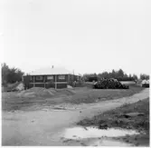 Vägstation E6, Mantorp, filial Slaka. Byggnad med garageanläggning i souterrängvåning. Upplag av bränslefat till höger.