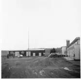 Vägstation E8, Ringarum. Byggnad med verkstadsdel, kontorsdel och garagedel. Personbilar parkerade framför garageport. Stående bränsletank vid gaveln. Förrådsbyggnad i fonden. Bensinpumpar (drivmedelspumpar), drivmedelstank, 