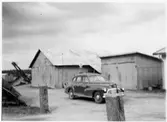Vägstation F6, Kvarnarp. Förrådsbyggnader av korrugerad plåt. Skopelevator och sidoplog i bakgrunden. Personbil framför byggnaderna. Bensinpump (drivmedelspump).
