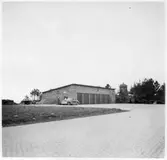 Vägstation H12, Bengtstorp. Garagebyggnad, av tegel. Bensinpumpar (drivmedelspumpar), två personbilar, mansperson. I bakgrunden en väderkvarn. Invid gatstensbelagd väg. Garageuppfarten med gatsten.