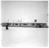 Vägstation M7, Dalby. Vägstationens huvudbyggnad, med från vänster verkstad, förråd, kontor. Bensinpump (drivmedelspump) på garageplan. Två personbilar framför kontoret. Tre manspersoner mellan bilarna.