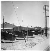 Vägstation M11, Södervidinge. Garagebyggnad, innanför nätstängsel. Uppställda spetsplogar. I bakgrunden bostadshus och spiran på ett kyrktorn.