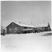 Vägstation M12, Marieholm. Förrådsbyggnad. I bakgrunden garagebyggnad. Framför byggnaden två lastbilar med två manspersoner som arbetar vid lastbilarna. Bensinpump (drivmedelspump). Vid gavel slipstenbock med slipsten.