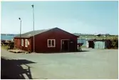 VFO, Färjevarvet, Fridhem. Förrådsbyggnad. Färja i vattnet framför byggnaden. Bostadshus i bakgrunden.