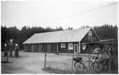 Vägstation P13, Brålanda. Garage- och förrådsbyggnad. Cykelställ med cyklar, bensinpumpar (drivmedelspumpar), sidoplogar, spetsplogar, personbil Volvo PV. Lågt nätstängsel.