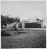 Vägstation R7, Götene. Vägstationens huvudbyggnad. Garagebyggnad (till vänster) hopbyggd med tvåvåningshus, eventuellt med bostad för vägmästaren i övervåningen, kontor i bottenvåningen.  Förrådsbyggnad i anslutning till garaget. Cementrör upplagda på lastkaj. Vid förrådsbyggnaden manskapsskåp för manskapets personliga utrustning och redskap.