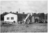 Vägstation R11, Grästorp. Garagebyggnad. Framför byggnaden lastningselevator (skopelevator), väghyvel, eventuell Svedala typ BD2. Smörjbrygga vid gaveln. Lösa hjul till väghållningsmaskiner.