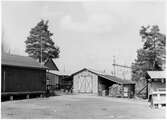 Vägstation T7, Frövi. Förrådsbyggnader, garagebyggnad i korrugerad plåt. Upplag av virke under sidotak. Järnväg (bangård) i bakgrunden.