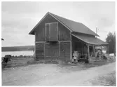 Vägstation W15, Särna. Förrådsbyggnad invid mindre väg. Särnasjön i bakgrunden.