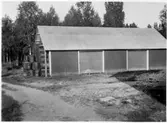 Vägstation Vmo X4, filial Freluga. Förrådsbyggnad, med upplag vid gaveln, bränslefat, tjärtunnor.