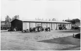 Vägstation Y6, Älandsbro. Förrådsbyggnad, eventuellt äldre garagebyggnad. Lastmaskiner och lastbil framför byggnaden. Bondgård i bakgrunden.