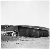 Vägstation Y12, Sollefteå. Förrådsbyggnad. Upplag av cementrör vid gaveln. Vägsladdar framför byggnaden.