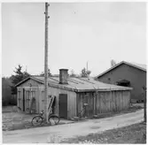 Y län. Före detta hamnförrådet. Förråds- och garagebyggnad, eventuellt också smedja,  vid infarten, med grind, till området. Cykel lutad mot telefonstolpe.