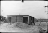Vägstation AC3, Umeå. Förrådsbyggnad. Upplag av sand, bränslefat. Skottkärra i förgrunden.