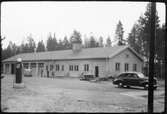 Vägstation AC4, Vännäs. Vägstationens huvudbyggnad, med från vänster garage, kontor, förråd och verkstad. På garageplan två personbilar, bensinpump (drivmedelspump), cyklar mot väggen, tre manspersoner.