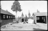 Vägstation AC8, Burträsk. Förrådsbyggnader och del av garagebyggnaden (vägstationens huvudbyggnad). Mansperson sittande på marken, lagande lastbilsdäck.