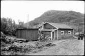 Vägstation AC21, Tärnaby, filial Umfors. Garage- och förrådsbyggnad, eventuellt även manskapsbod (baksidan). Upplag av diverse materiel. Personbil och grupp av manspersoner vid vägen.