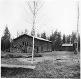 Vägstation BD2, Älvsbyn, filial Vidsel. Garage- och förrådsbyggnad. Förrådsbyggnad i bakgrunden. Bensinpump  (drivmedelspump). Vedupplag vid gaveln.