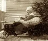 Robert Nelson (född 1908) sittandes i en leksaksbil utanför en byggnad i USA, okänt årtal. Son till Carl Nelson (född i Sverige, död i USA) och Alma Nelson (född 1881 i Kållered - död 1959 i USA). Hennes far var Karl Eriksson från Vommedal Östergård 