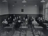Skolklass med pojkar. Södra eller norra folkskolan. Två elever står vid väggbröstningen längst bak, en pojke står mellan två bänkrader och övriga sitter med uppslagna böcker i sina bänkar. I taket hänger s k skomakarlampor.