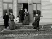 Tre män, två kvinnor och en hund på trappan till Himle härads tingshus. Ena dörrbladet står öppet och damen som står där bär en pälsstola.