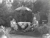Borgmästare Johan Janssen sitter tillsammans med frun Annie (f Ullgren) i sin lummiga trädgård under ett parasoll i kv Komedianten 1. Annie handarbetar. Spaljerade luktärter blomstrar i bakgrunden och i pelargoner står i urnorna vid sandgången.