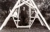 Två kvinnor sitter i en gunga/hammock, en man står vid sidan om.