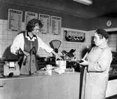 En kund står vid kassan och väntar på att personalen skall paketera hennes varor. Konsum Sörhaga, Viktoriagatan.
Butiken öppnade 1939.