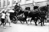 Hästvagn med två hästar och kusk samt medpassagerare, i uniform, rullar fram över en gata. Vagndelen bakom kuskens sittplats är klotformad. Den så kallade Kugleposten passerar kv Gustav på Torggatan mot norr. 
Närmst kameran två flickor och på husets fasad bakom vagnen syns skyltar med texten 