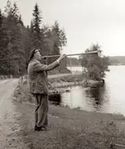 En man står och blåser i en slags tuta vid en sjö i Värmland år 1937. Elever och lärare från Kållered är på skolresa. Skolresan gick genom Dalsland/Värmland till den norska gränsen, och anordnades av Daga och Gunnar Alander. Gunnar var folkskollärare i Kålleredsskolan. 
Relaterade motiv: A3101, A3103 - A3108.