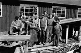 Gruppbild från Alingsås Snickerifabrik, kv Gjutaren. 11 män står/sitter framför en husvägg. Runt fötterna ligger bråte och olika träbitar.