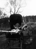 Två små barn, den ena sitter i en vagn medan den andra står bredvid.