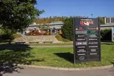 Liten park i Lindome centrum den 4 oktober 2016. I bakgrunden ses Lindome Spel & Tobak samt Lindomefloristerna i huset Almåsgången 2. I parken finns sittplatser samt en stor skylt som anger vilka affärsrörelser med mera som finns i centrum. Vy mot norr.