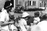 En kvinnlig sköterska och fyra små barn sitter utomhus på en gräsmatta, Sagåsens vårdhem cirka 1965 - 1970.