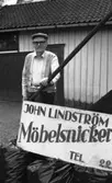 John Lindström (född 1902 i Lindome, död 1989 i Kållered) står utanför sitt snickeri på Gamla Riksvägen 79 (nu: Gamla Riksvägen 81), 1980-tal. På skylten står det 