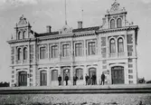 Falkenbergs järnvägsstation. Stationen tillkom i samband med Mellersta Hallands järnväg som invigdes 1886 och gick mellan Varberg-Halmstad. På perrongen står sex män som ser ut att vara uniformerade såsom stationspersonal. I modern tid har järnvägen dragits öster om stadens centrum och sträckan med ny station invigdes 15 juni 2008.