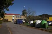Återvinningskärl för glas vid parkeringsplatsen i Lindome centrum, den 4 oktober 2016. I bakgrunden ses vårdcentralen och Apoteket i byggnaden Almåsgången 7. Vy mot norr.
