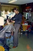 Frisören och ägaren Leif Westerberg klipper en kund (okänd kvinna) som sitter i en stol, Hårmarkarna på Våmmedalsvägen 10 i Kållered 1980-tal.