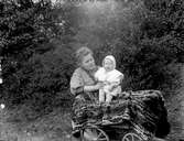 Kvinna och litet barn vid en vagn i en trädgård. Kvinnan på bilden är troligtvis bekant till fotografen.