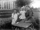 Kvinna med två små barn i trädgården framför fotoateljén på Mogärde i Vetlanda. Kvinnan är troligtvis fotografens syster, Amy Ekström och barnen är troligtvis hennes döttrar, Anna Karin och Inga Kristina.