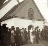 Människor står samlade utanför Kållereds kyrka när brudparet Iris Johansson (1913 - 2004, gift Persson), Livered och John Persson (1909 - 1982) vandrar ut, 1938-10-23. Bruden bär Kållered församlings brudkrona, som används för första gången. 
Det var dubbelbröllop denna dag då Iris kusin Iréne Karlsson (1912 - 2016, gift Karlsson), Tållered gifte sig med Erik Karlsson (1912 - 1997). Relaterade motiv: A3099 och A3102.