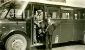 En chaufför och en konduktör (namnppgifter saknas) står i/vid en buss tillhörande Kållered bussbolag (Kålleredbussarna), 1930-tal. På bussen står det Göteborg-Anderstorp. Fordonet står troligtvis utanför garaget på Gamla Riksvägen/Streteredsvägen.