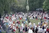 Barn och vuxna dansar kring stången vid midsommarfirande på Ekensås cirka 1982. Spelmannen Frank Efraimsson (i blå skjorta och vita byxor ståendes vid midsommarstången) spelar dragspel.