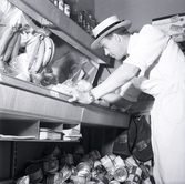 En man i hatt och vit rock vid varor i en konsumbutik.