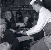 En man i vit rock som håller i en bricka med en godsak bland människor i Konsum.