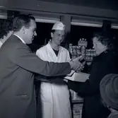 En man i kostym som skakar hand med en kvinna som håller i ett paket i Konsum. En man i vit båtmössa och vit rock är centrerad.